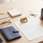 Inquadratura dall’alto di un tavolo in legno chiaro con un taccuino blu scuro, un piccolo raccoglitore blu navy, occhiali, una busta di carta naturale, un blocco per appunti, chiavi, un foglio con schemi astratti senza testo sormontato da un fermacarte dorato e una tazza grigia su un sottobicchiere.