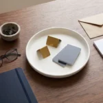 Inquadratura dall’alto di una scrivania in legno scuro con una cartella e un taccuino blu, occhiali, una ciotolina con fermagli, una busta e un blocchetto di carta, una tazza scura e, al centro, un piatto bianco con piccoli divisori dorati e tessere grigie senza testo.
