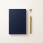 Inquadratura zenitale su una superficie beige calda con un taccuino blu navy chiuso, una penna in ottone lucido e un piccolo accento dorato.