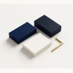 Composizione geometrica dall’alto su superficie bianca calda con tre blocchi rettangolari lisci nei toni del blu navy, blu scuro e bianco caldo, affiancati da un elemento angolare minimalista in ottone dorato opaco, senza scritte né loghi.