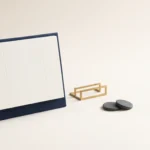 Composizione concettuale su superficie chiara con un supporto da tavolo blu scuro contenente una scheda con griglia astratta senza numeri né testo, affiancato da un piccolo fermacarte geometrico dorato e due pietre lisce color carbone.