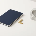 Inquadratura zenitale su una superficie chiara con un taccuino blu scuro chiuso, un piccolo marcatore geometrico in ottone dorato e un blocco ceramico astratto.