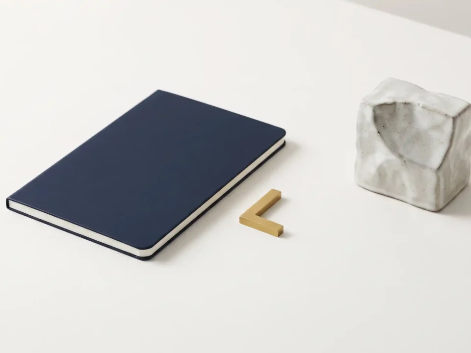 Inquadratura zenitale su una superficie chiara con un taccuino blu scuro chiuso, un piccolo marcatore geometrico in ottone dorato e un blocco ceramico astratto.