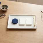 Inquadratura dall’alto di una scrivania in legno chiaro con una cartella e un taccuino blu, occhiali, una ciotolina con fermagli, una busta e un blocco di carta, una tazza scura e, al centro, un vassoio bianco con un elemento circolare blu e due rettangolari bianchi affiancati da un dettaglio dorato, senza testo né loghi.